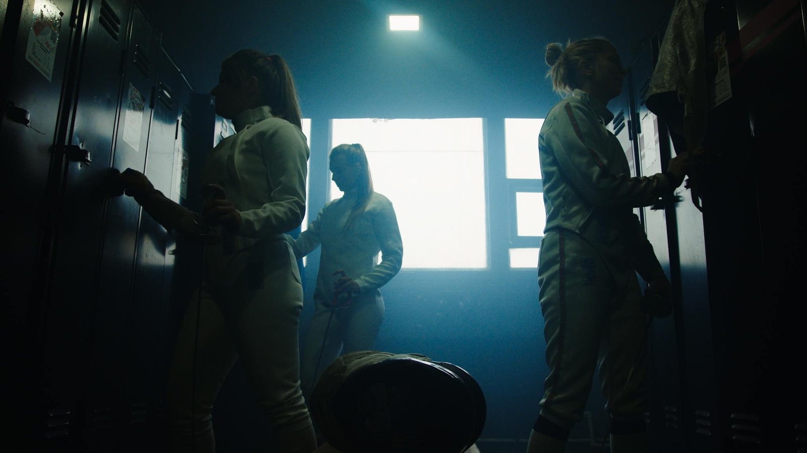 Three female fencers getting ready in a locker room, backlit by window light.